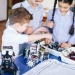 Excited little kid and two smiling teen friends construct robotic toys while playing during their leisure time in technical workshop. Intelligent activity.